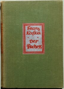 Capa de tecido verde da primeira edição do livro O Processo, de Franz Kafka, publicada em 1925, com uma etiqueta central contendo o título impresso em letras vermelhas de estilo expressivo.